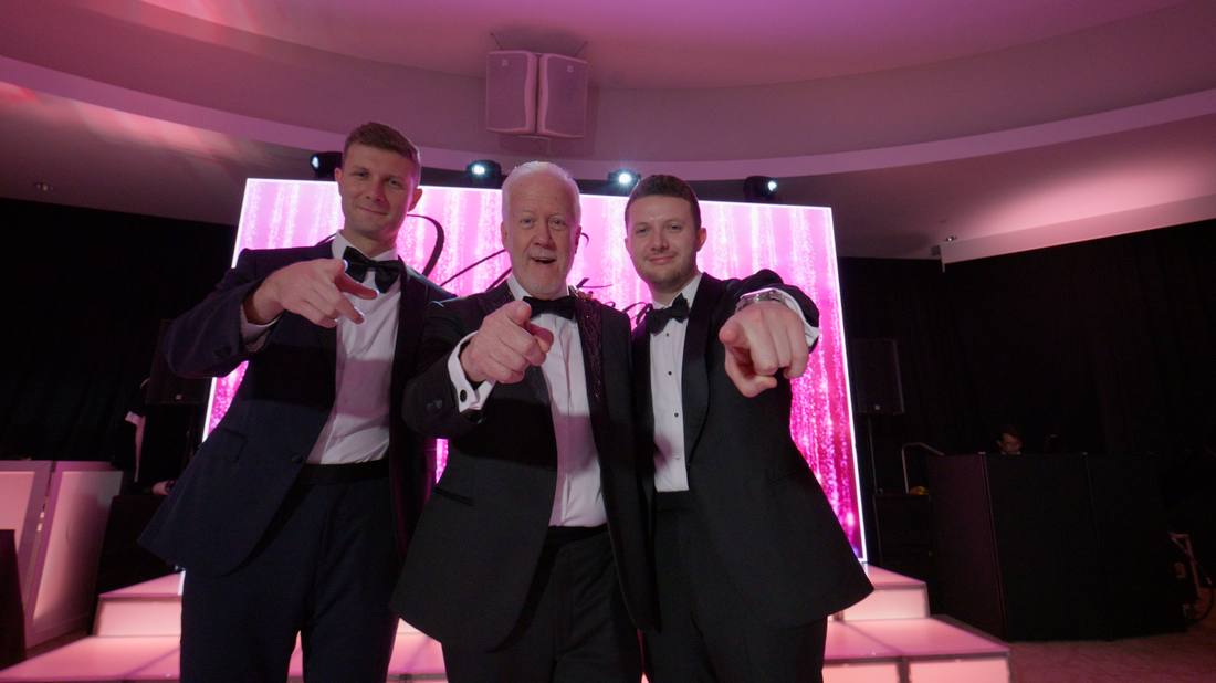 Three men in tuxedos posing in front of a glittering pink stage backdrop